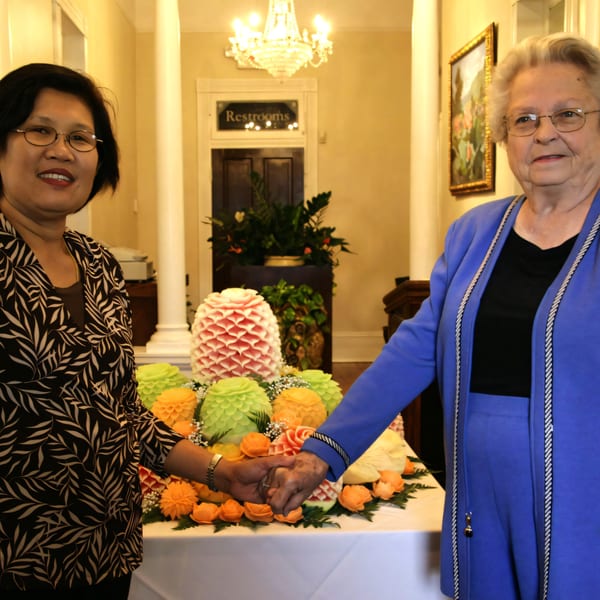 Two women holding hands over a table adorned with Thai fruit carvings, including a pineapple.