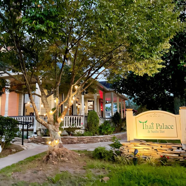Exterior of Thai Palace & Sushi Bar at dusk, with a lit sign and string lights on a tree.