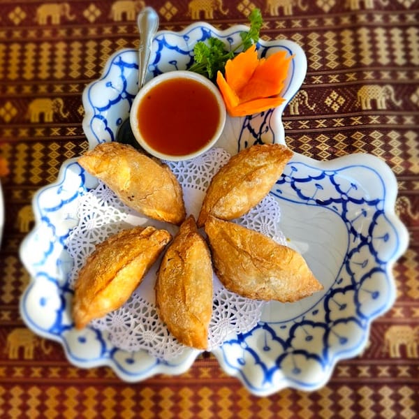 Six golden-brown Thai curry puffs arranged around a small bowl of dipping sauce on a decorative plate.